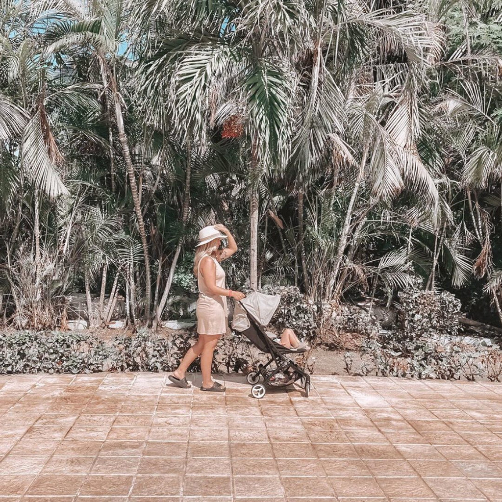 Mom pushing a toddler in a forward-facing gently open box stroller in front of palm trees
